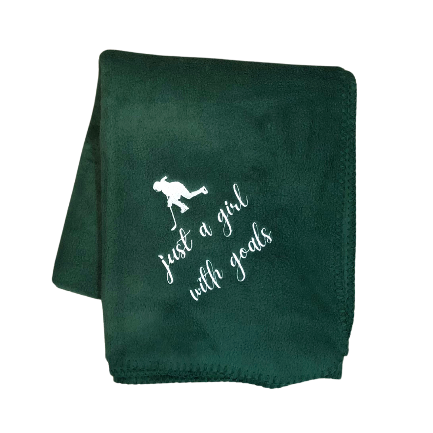 A green fleece blanket with hockey-themed embroidery and the phrase 'just a girl with goals' stitched in white