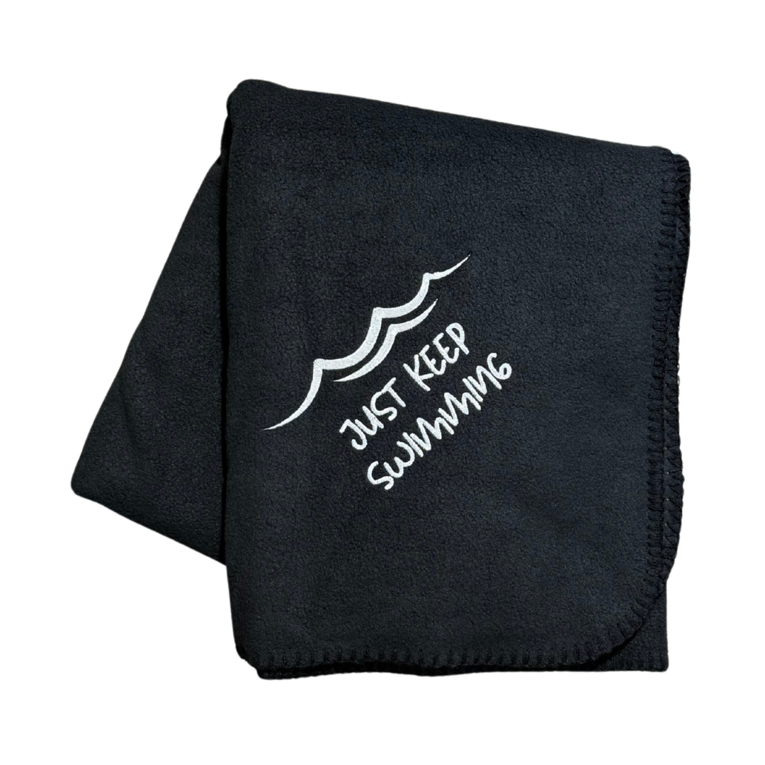 Black blanket with 'Just Keep Swimming' text and wave design in white embroidery