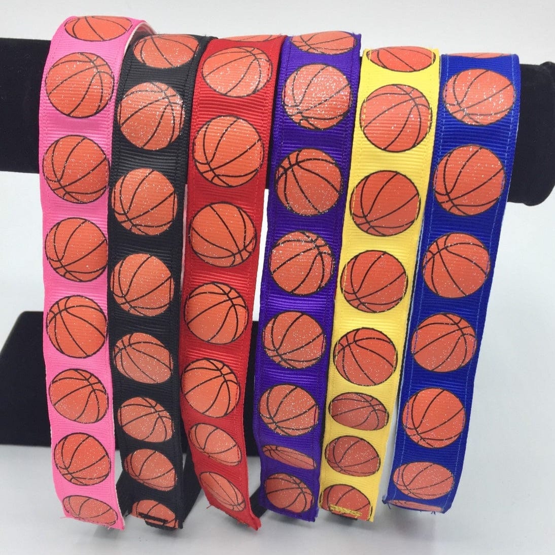 six basketball headbands showing all the colors available. Each has orange glittery basketballs. The colors shown are pink, black, red, purple, gold, and royal blue.