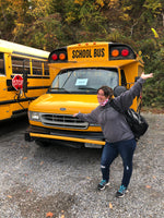 Maggie Eastman posing excitedly in front of short yellow school bus that she purchased in Pittsburgh PA