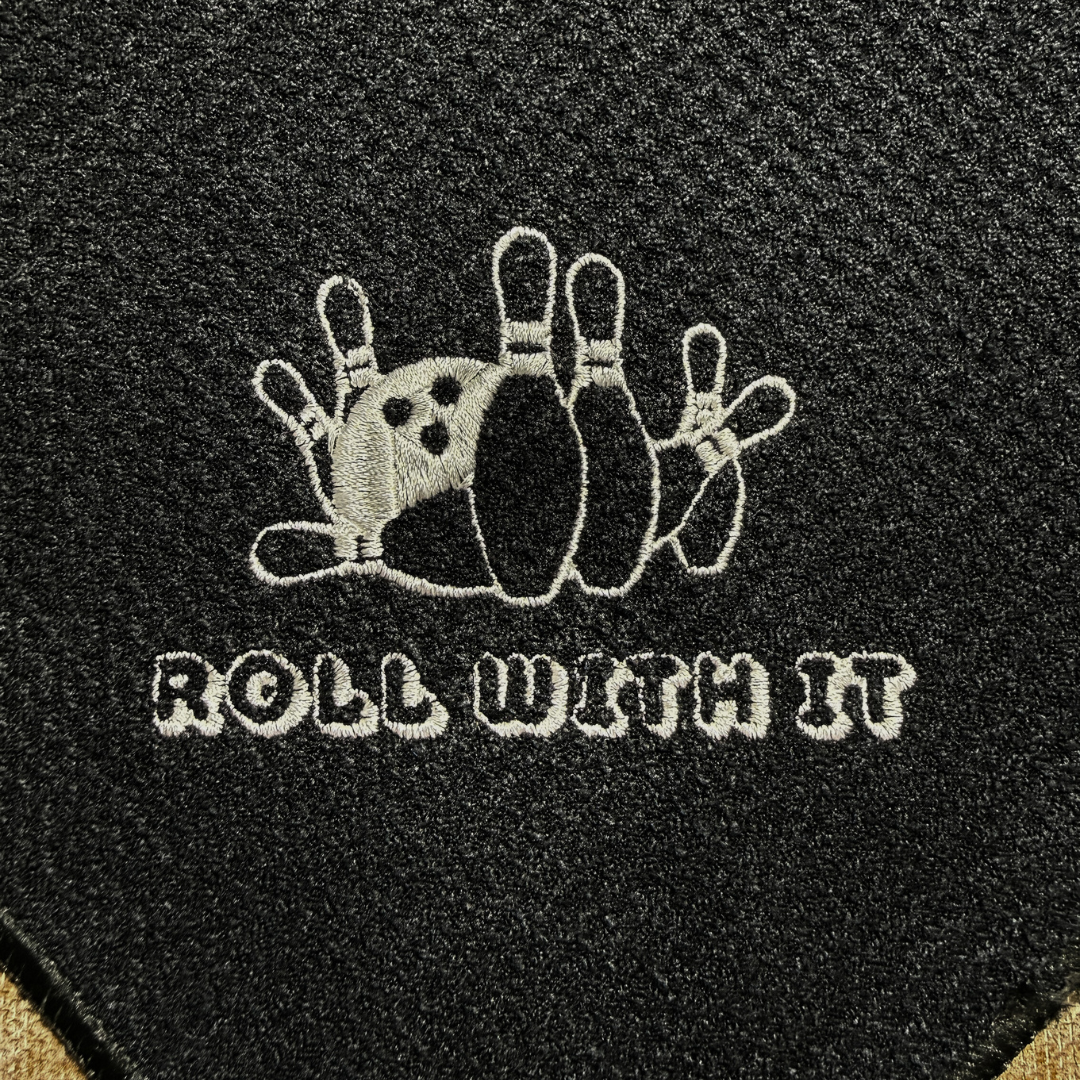 Black bowling ball with white embroidered design and 'Roll with It' text on a black background