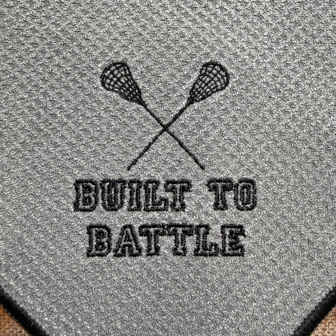 Embroidered design of crossed lacrosse sticks with 'Built to Battle' text on a textured gray towel.