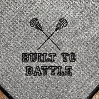Embroidered design of crossed lacrosse sticks with 'Built to Battle' text on a textured gray towel.