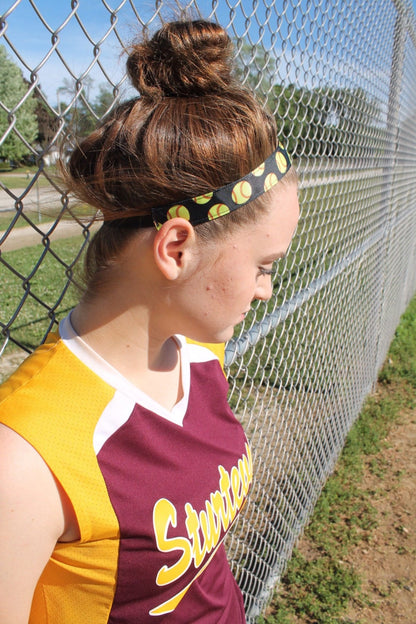 girl leaning on a fence wearing a softball jersey and black softball headband with her hair in a topknot